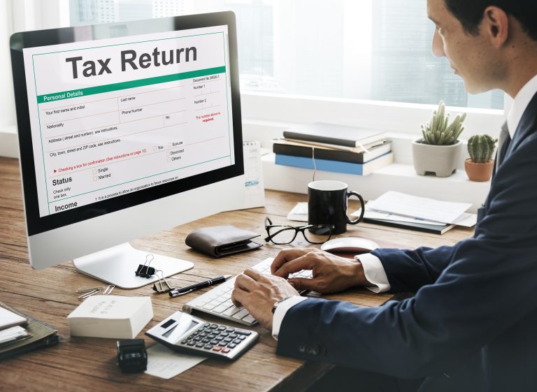 A person holding a calculator over tax forms, with a coin stack and a refund cheque beside a laptop, representing income tax